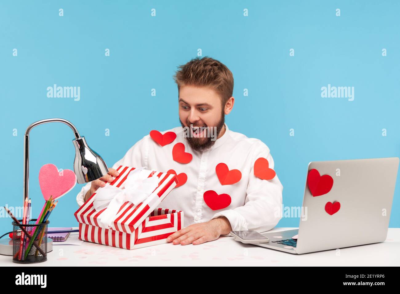 Glücklich überrascht Mann mit Bart in weißem Hemd in roten klebrigen Herzen Unboxing Gegenwart und Blick nach innen mit Lächeln, zufrieden mit Geschenk. Studio im Innenbereich Stockfoto