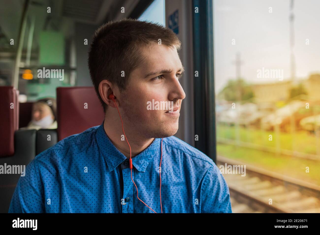 Ein junger europäer in einem blauen Hemd, der Musik auf Kopfhörern hört, schaut aus dem Fenster, während er in einem modernen Elektrozug unterwegs ist. Stockfoto