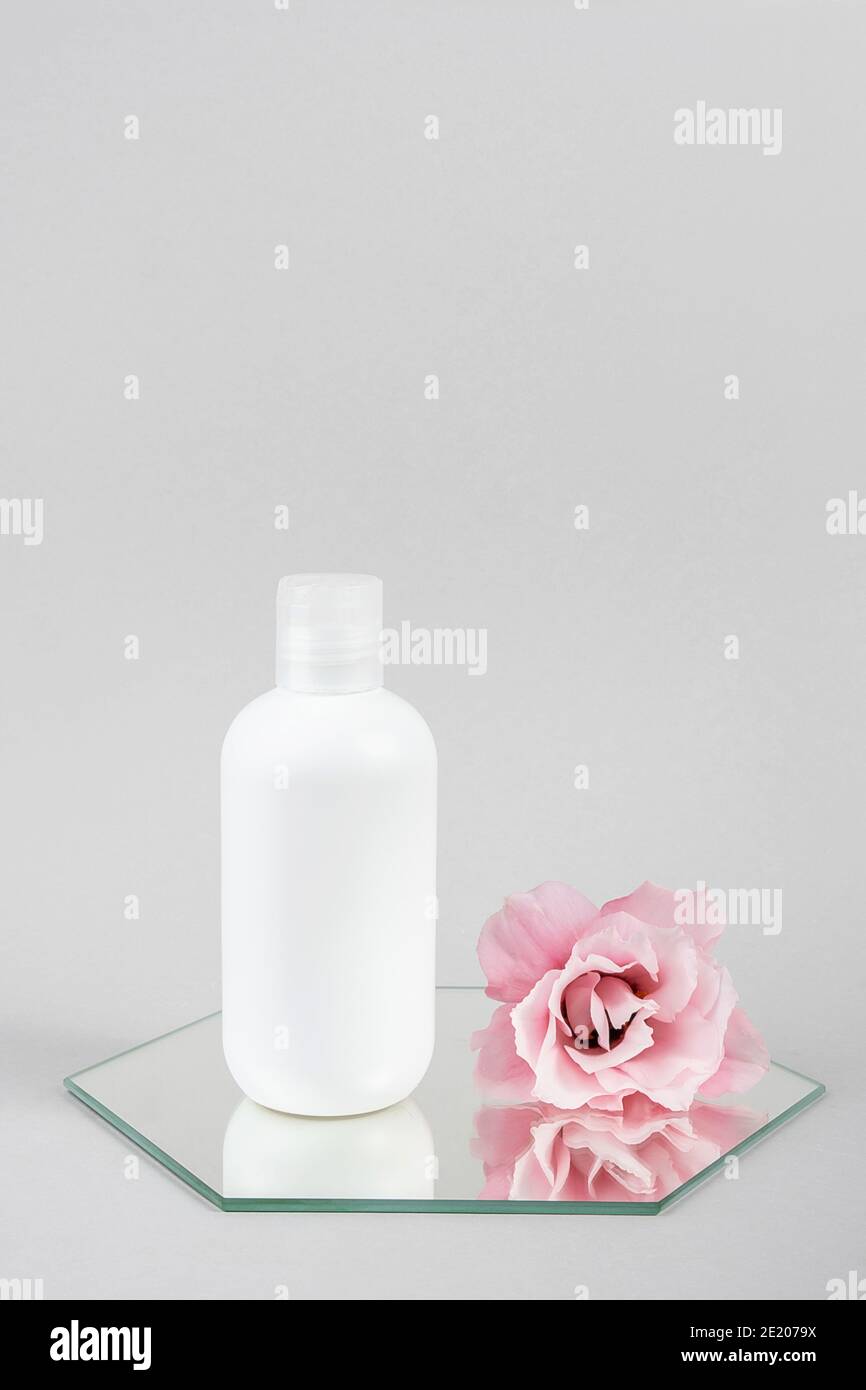 Weiße kosmetische leere Flasche und rosa Blume auf Spiegel, grauer Hintergrund. Natural Organic Spa Kosmetik Schönheitskonzept Mockup, Vorderansicht. Stockfoto