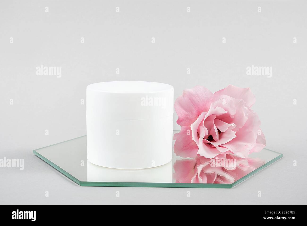 Weiße kosmetische leeres Glas und rosa Blume auf Spiegel, grauer Hintergrund. Natural Organic Spa Kosmetik Schönheitskonzept Mockup, Vorderansicht. Stockfoto