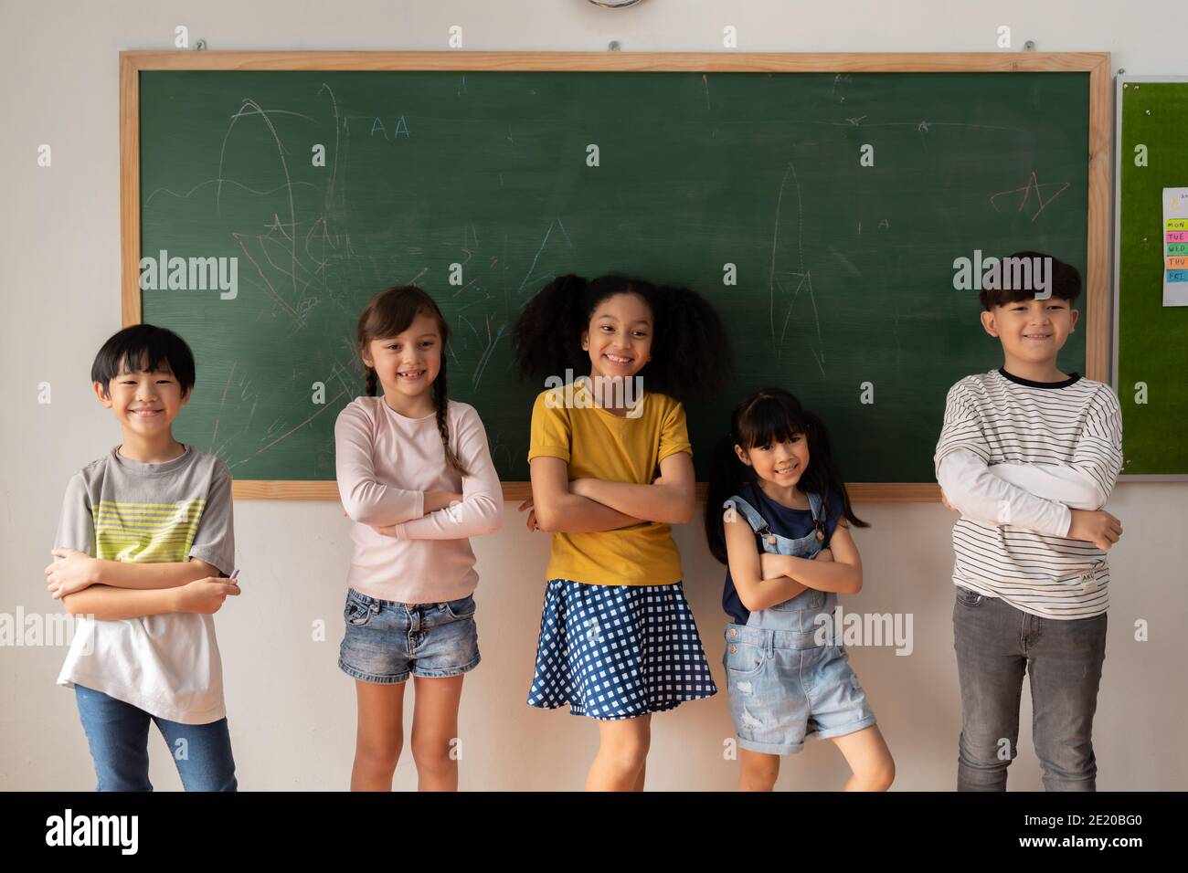 Glückliche multiethnische Kinder in lässiger Kleidung, die Arme kreuzt und lächelt Während des Unterrichts in der Grundschule in der Nähe der Tafel stehen Stockfoto