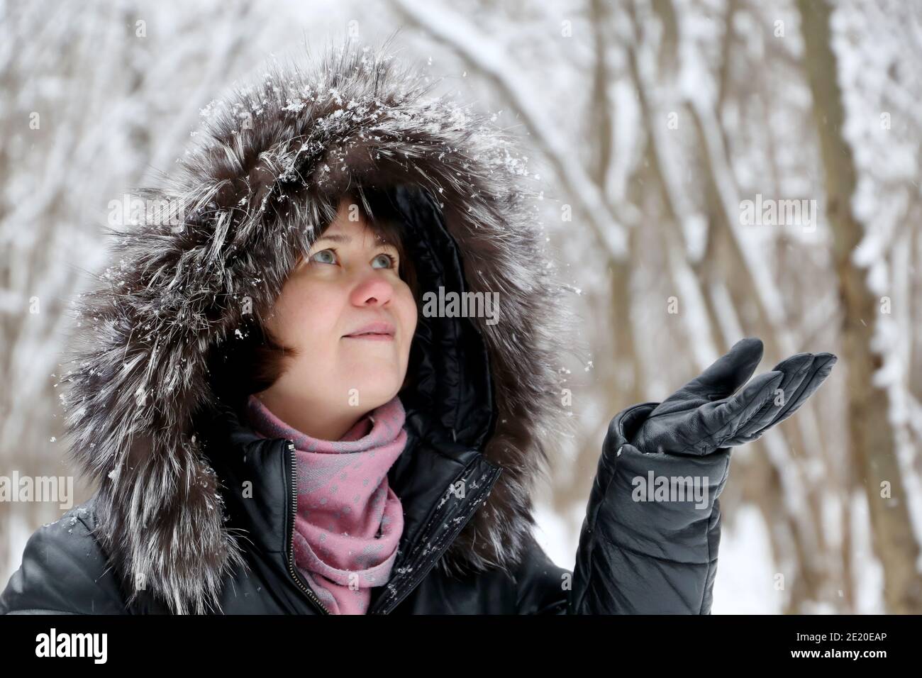 Glückliche Frau in Pelzhaube genießen das verschneite Wetter, fängt Schneeflocken mit ihren Händen. Schneefall im Winterpark, kalte Jahreszeit Stockfoto