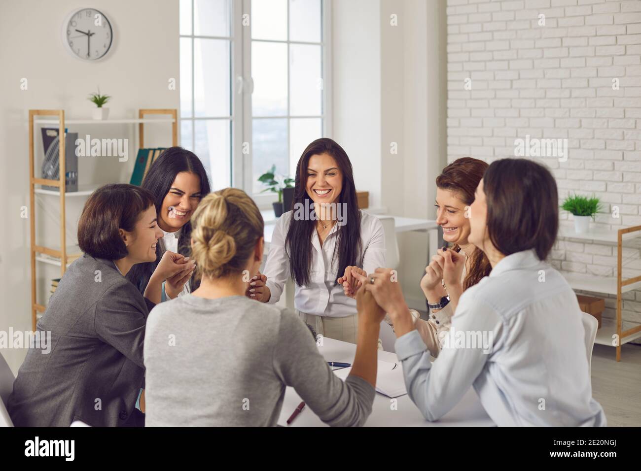 Team von glücklichen jungen Geschäftsfrauen, die am Bürotisch sitzen Und Hände halten Stockfoto