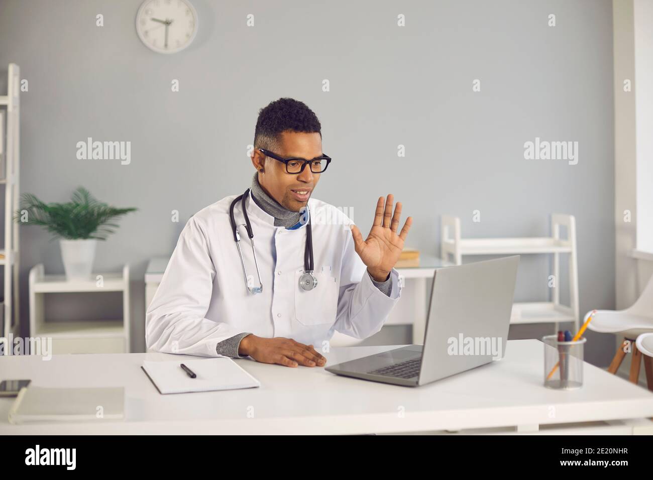 Online-Arzt sitzt am Schreibtisch, Video ruft seinen Patienten und winkt Hand an Laptop Stockfoto