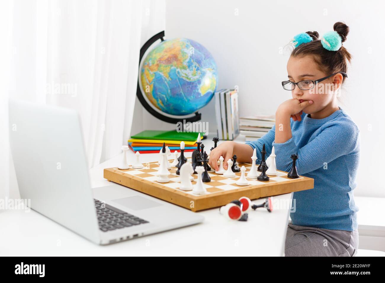 Portrait kaukasisches kleines Mädchen spielt Schach am Tisch im Innenraum mit Schachbrett. Kleines Mädchen lernt Schach auf einem Laptop online Stockfoto