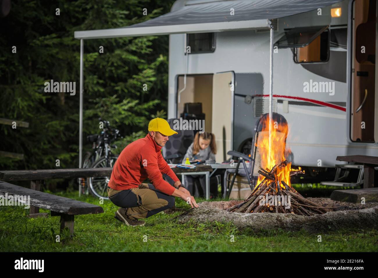 Kaukasische Familie auf dem Sommercampingplatz. Vater Verbringt Zeit Am Lagerfeuer. Seine Tochter schreibt Urlaub Destination Letter neben RV Camper Van. Stockfoto