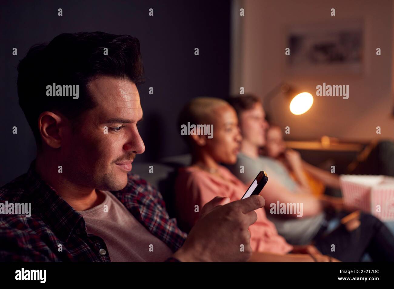 Mann mit Mobiltelefon, während Freunde zu Hause Fernsehen Abends Stockfoto