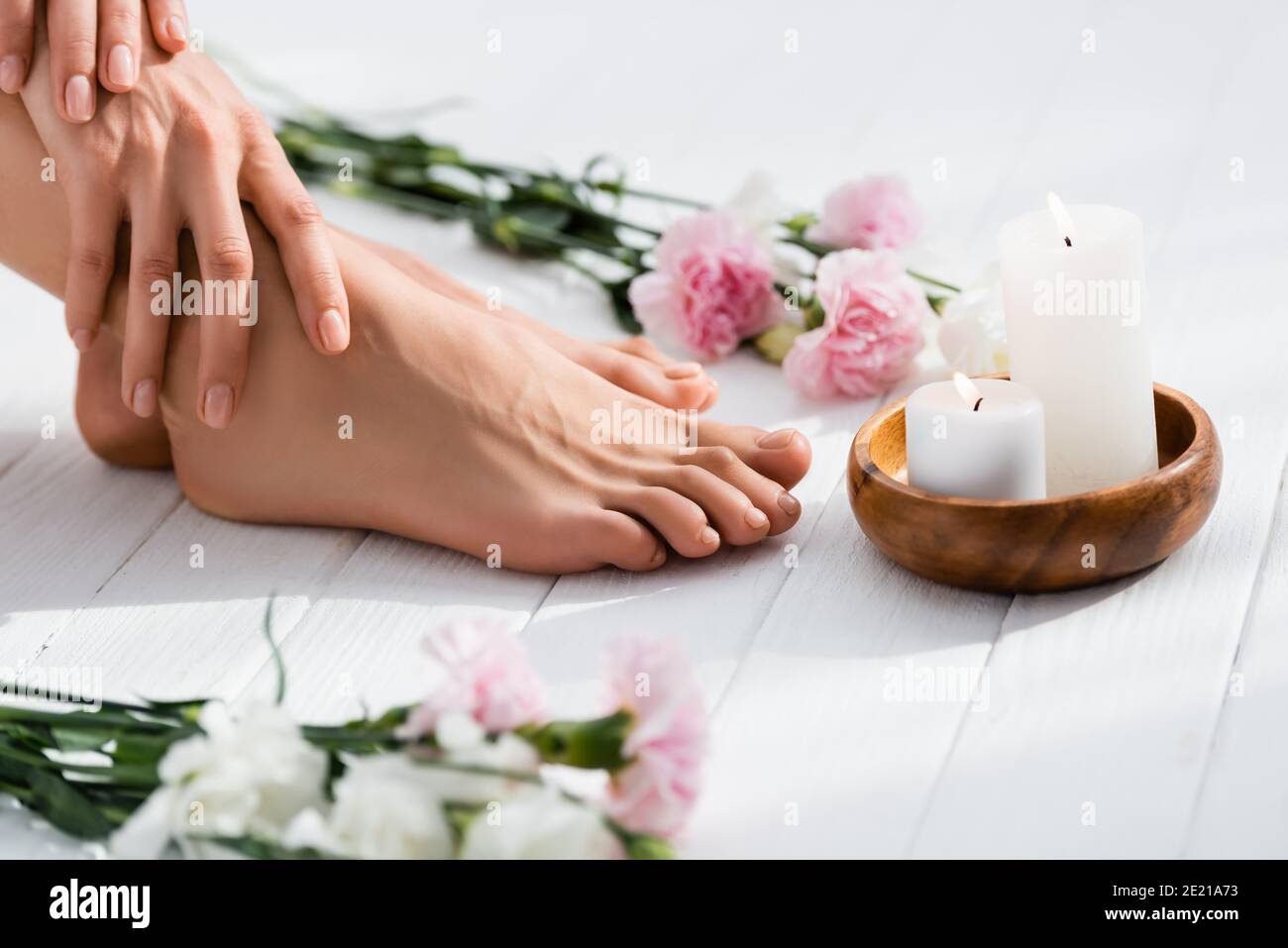 Beschnittene Ansicht der Frau mit gepflegten Händen und Füßen in der Nähe von Kerzen und Blumen auf weißer Holzoberfläche, verschwommener Vordergrund Stockfoto