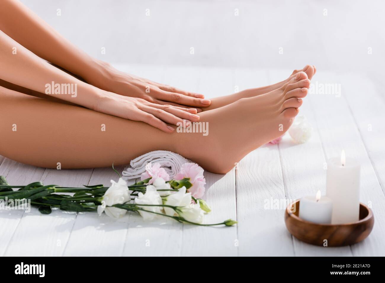 Beschnittene Ansicht der Frau mit gepflegten Händen und Füßen in der Nähe Weiße eustoma Blumen und Kerzen auf weißer Holzoberfläche Stockfoto
