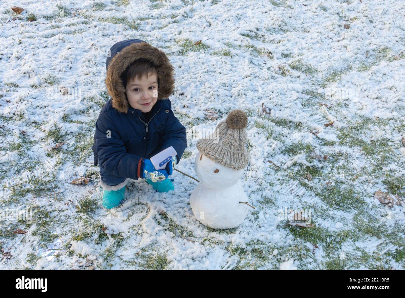 Kleiner Junge in einem Mantel, Handschuhen und blauen Stiefeln, die an einem Wintertag mit seinem Schneemann hinunterhocken Stockfoto
