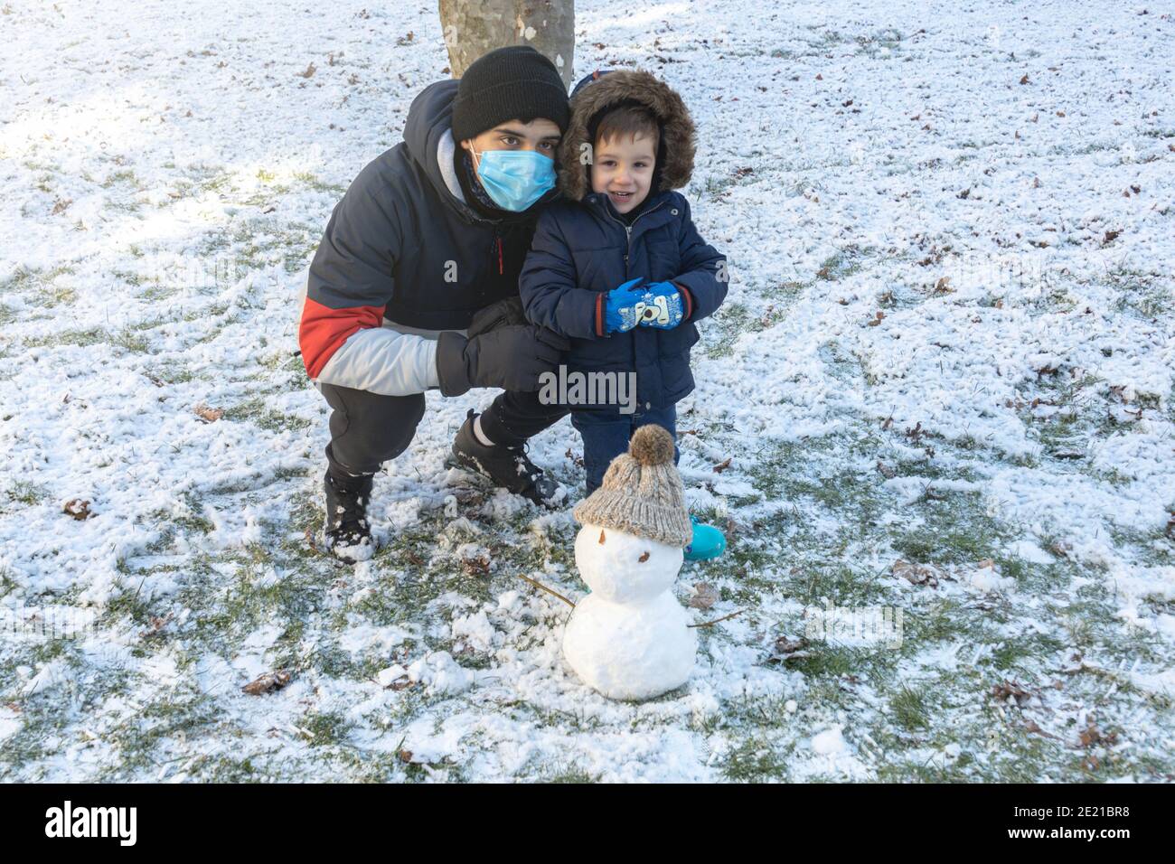 Teenager und Kleinkind in Schneemänteln, Handschuhen und Stiefeln mit ihrem Schneemann an einem kalten Wintertag Stockfoto