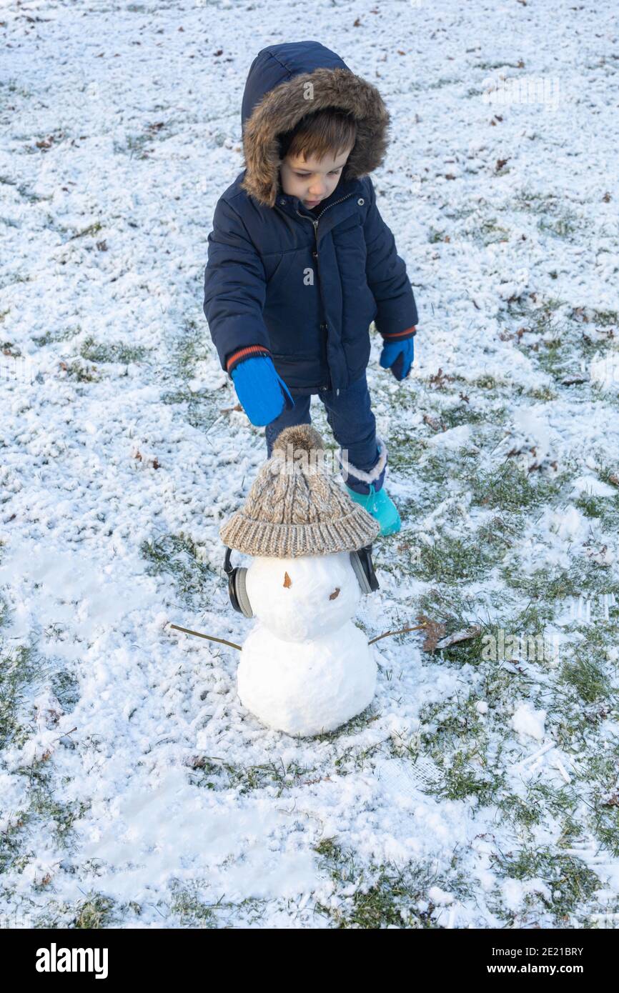 Kleiner Junge mit Mantel, Handschuhen und blauen Stiefeln zusammen mit seinem Schneemann, der an einem Wintertag einen Hut und Kopfhörer hat Stockfoto