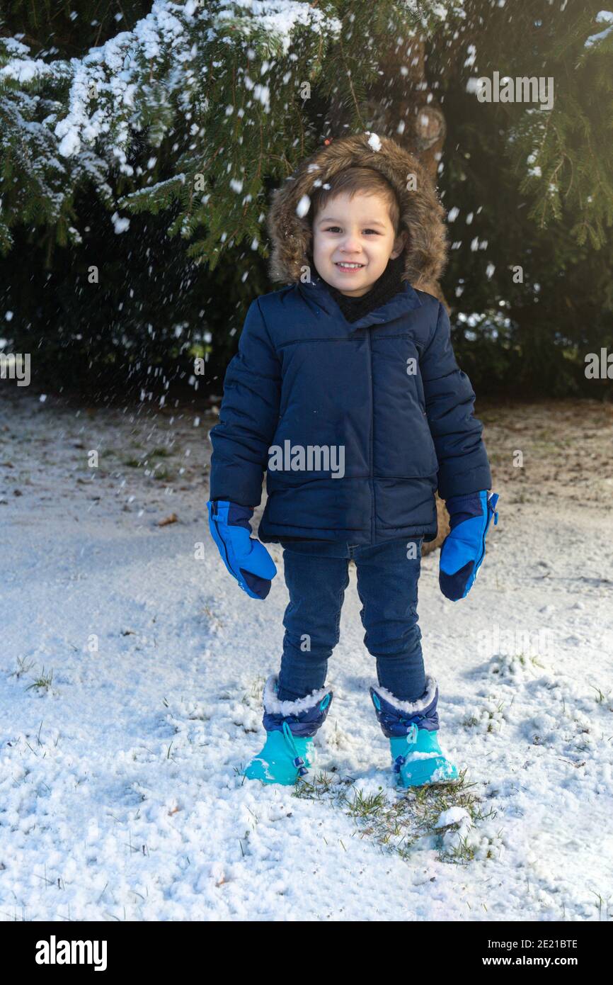 Kleines Kind in Mantel, Stiefel und blauen Handschuhen lächelnd, wie Schneeflocken fallen an einem Wintertag Stockfoto