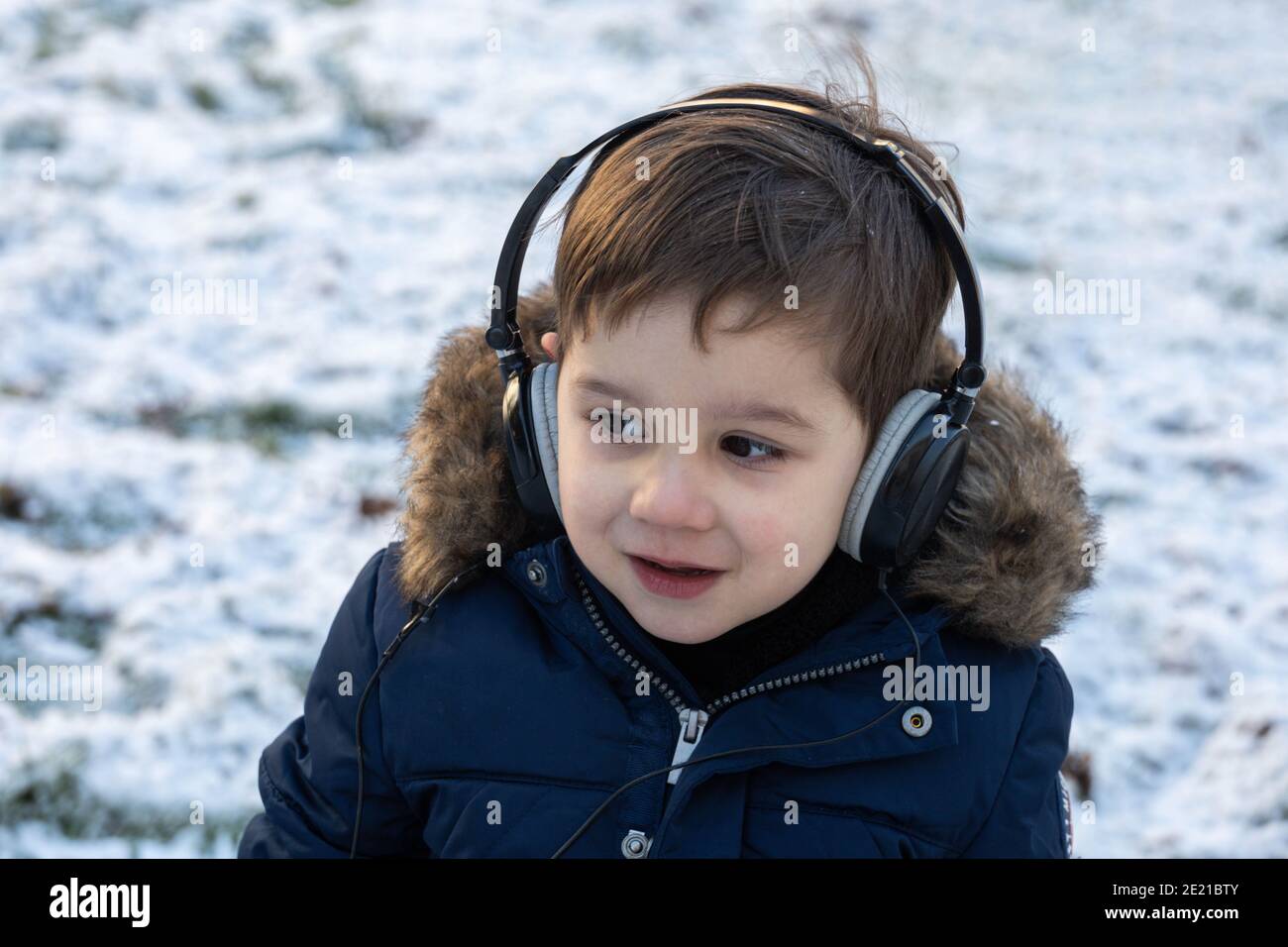 Selektiver Fokus eines geschützten Kleinkindes, das Musik mit hört Kopfhörer und Lächeln umgeben von Schnee an einem Wintertag Stockfoto