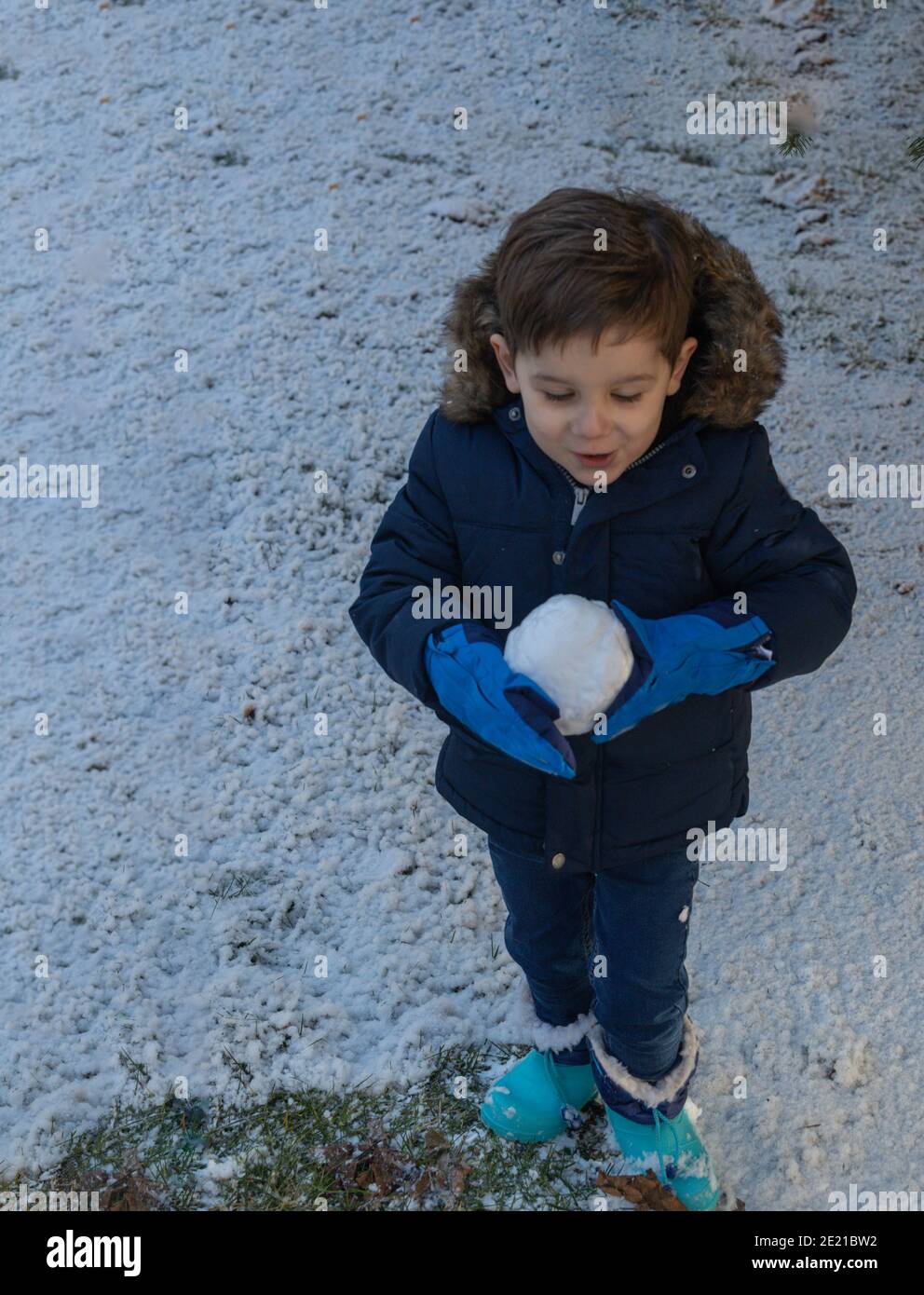 Kleinkind trägt blaue Stiefel und Handschuhe mit einem großen Schneeball An einem Wintertag Stockfoto