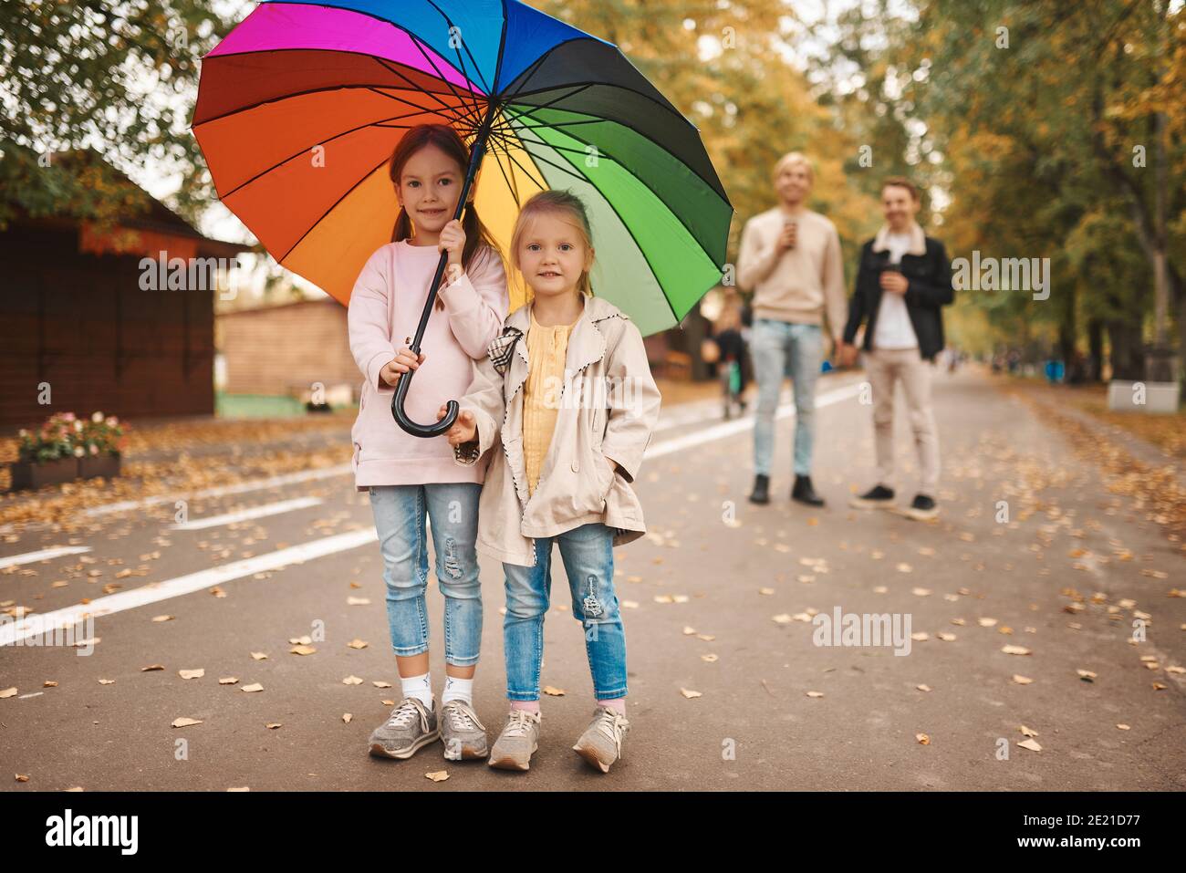 Zwei schwule Eltern mit ihren adoptierten Töchtern, die gemeinsam im Park spazieren. Zwei niedliche Mädchen stehen auf Vordergrund mit Regenbogen Regenschirm. Glückliche LGBT-Familie Stockfoto