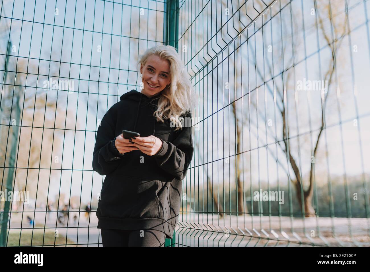 Glückliche hübsche Dame mit Handy in den Händen zu suchen Kamera während des Aufenthalts auf dem Basketballplatz Stockfoto