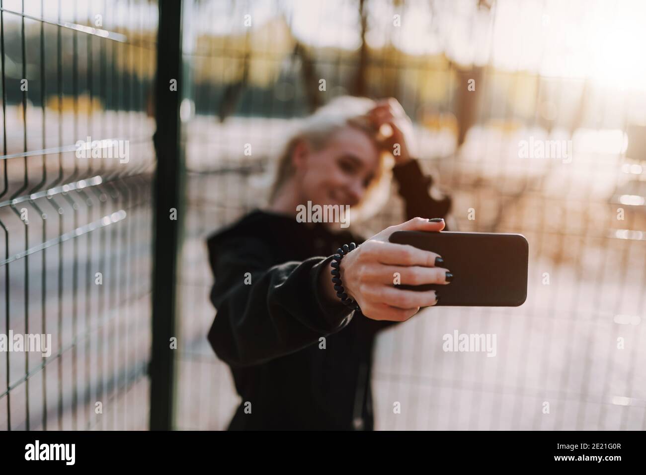 Lächelnde hübsche Dame hält Smartphone, während Selfie gegen Metall Gitter im Freien Stockfoto