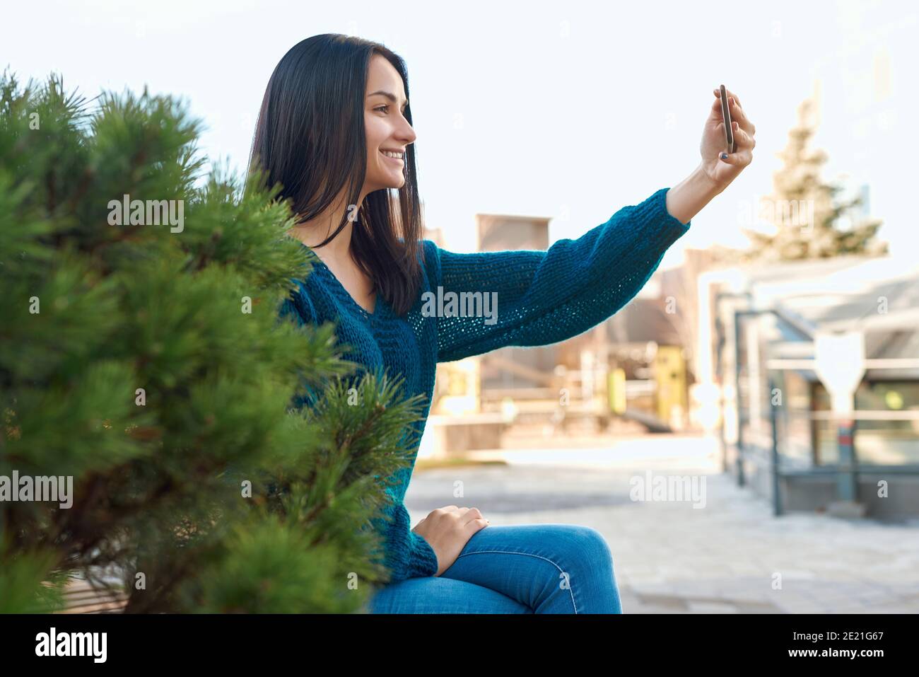 Seitenansicht einer lächelnden jungen Frau, die ein Selfie aufschießt Ihr Handy in der Nähe einer schönen dicken Zwergkiefer Stockfoto