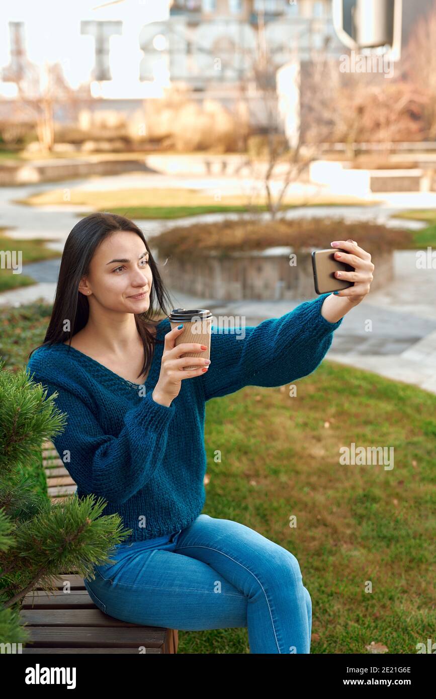 Positive Studentin macht Foto von sich selbst auf einem Handy Telefon während der Entspannung mit einer Tasse Kaffee in einer ruhigen Ecke eines malerischen Platzes Stockfoto