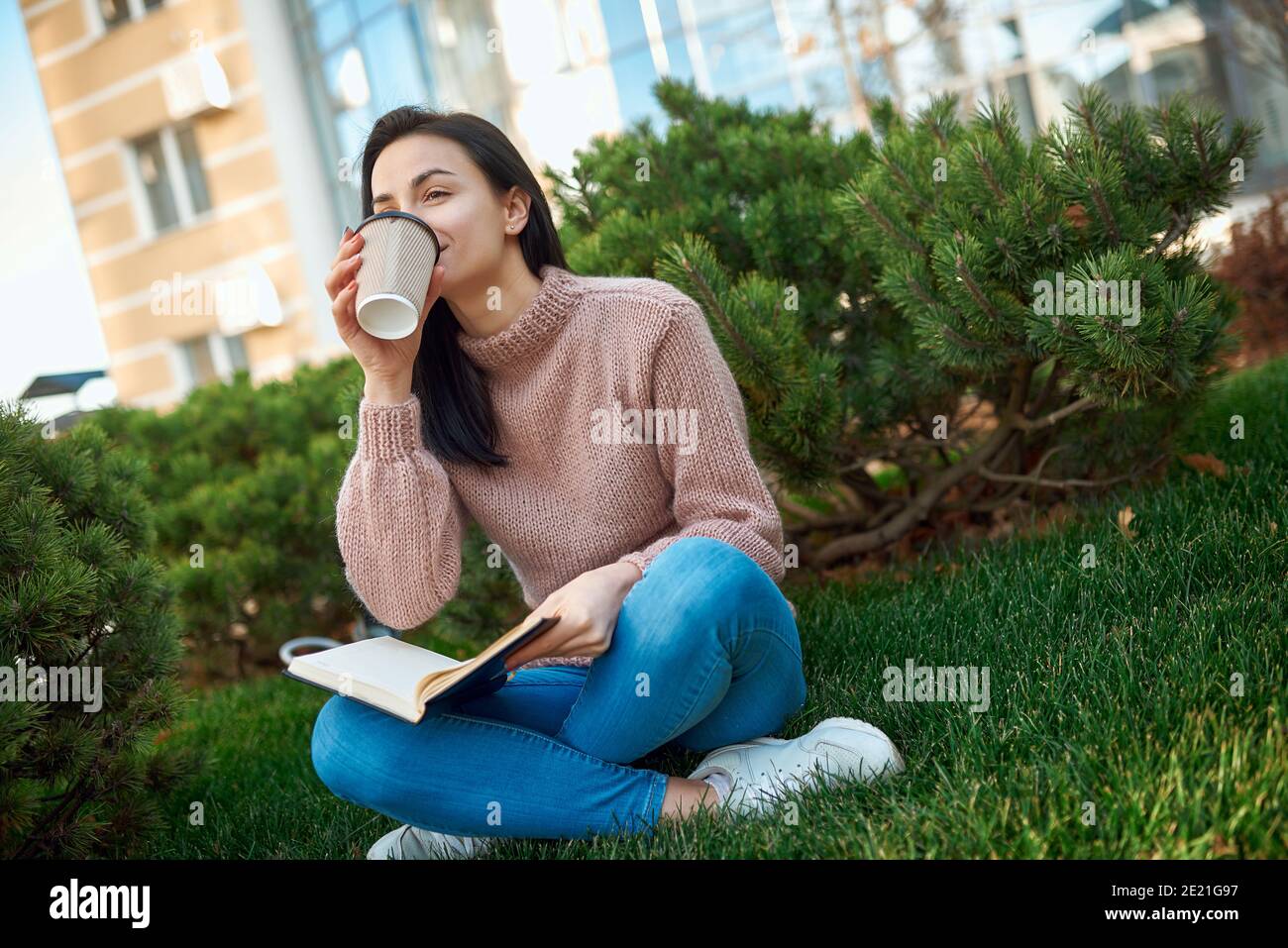 Hübsche junge Frau mit einem dicken daybook auf den Knien Genießen Sie heiße Getränke auf einem grünen Rasen in der Nähe hoch modern Gebäude Stockfoto