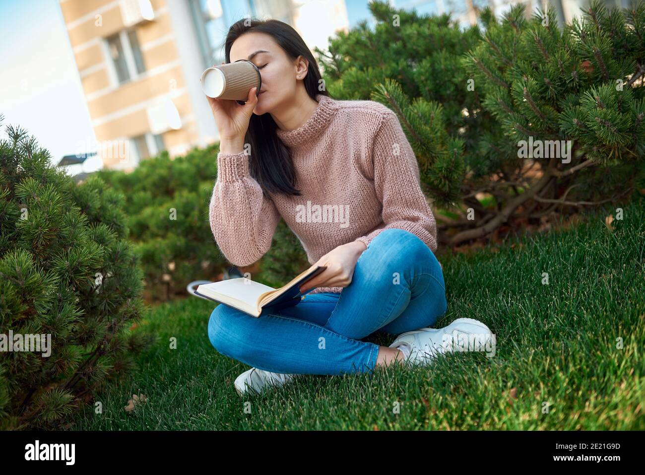Charmante junge Dame trinken Latte aus einem Papierglas und Setzen Sie das Copybook, während Sie auf grünem Gras innen sitzen Ein Türgarten Stockfoto