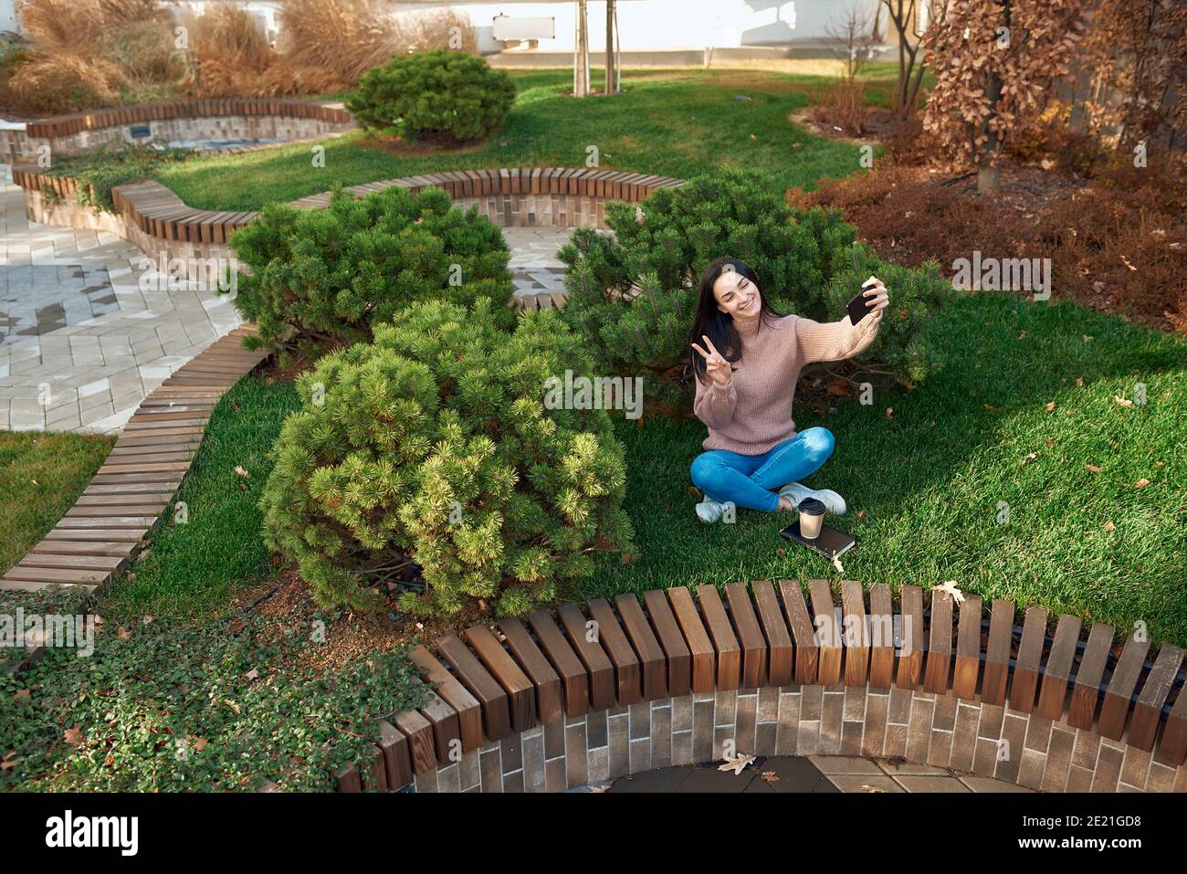 Fröhliche junge Frau schießen Selbstbild auf ihrem Telefon in Ein Stadtplatz mit schönen Kiefern im Hintergrund Stockfoto