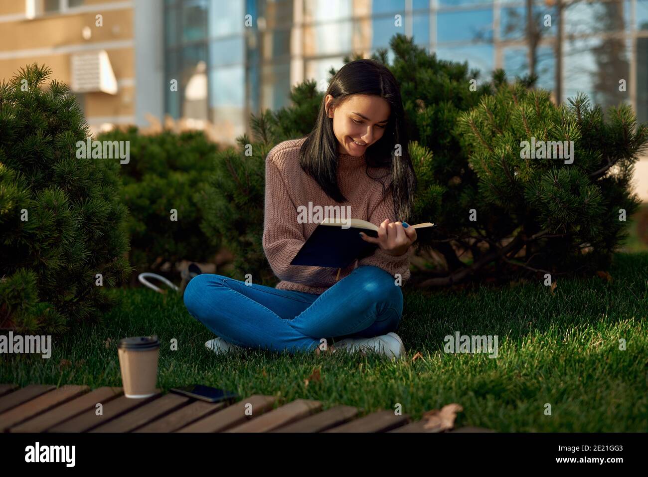 Schön aussehende lächelnde junge Modell posiert mit einem Buch auf Gras In ruhiger und friedlicher Hinterhof mit Kiefern außerhalb der modernen Gebäude Stockfoto