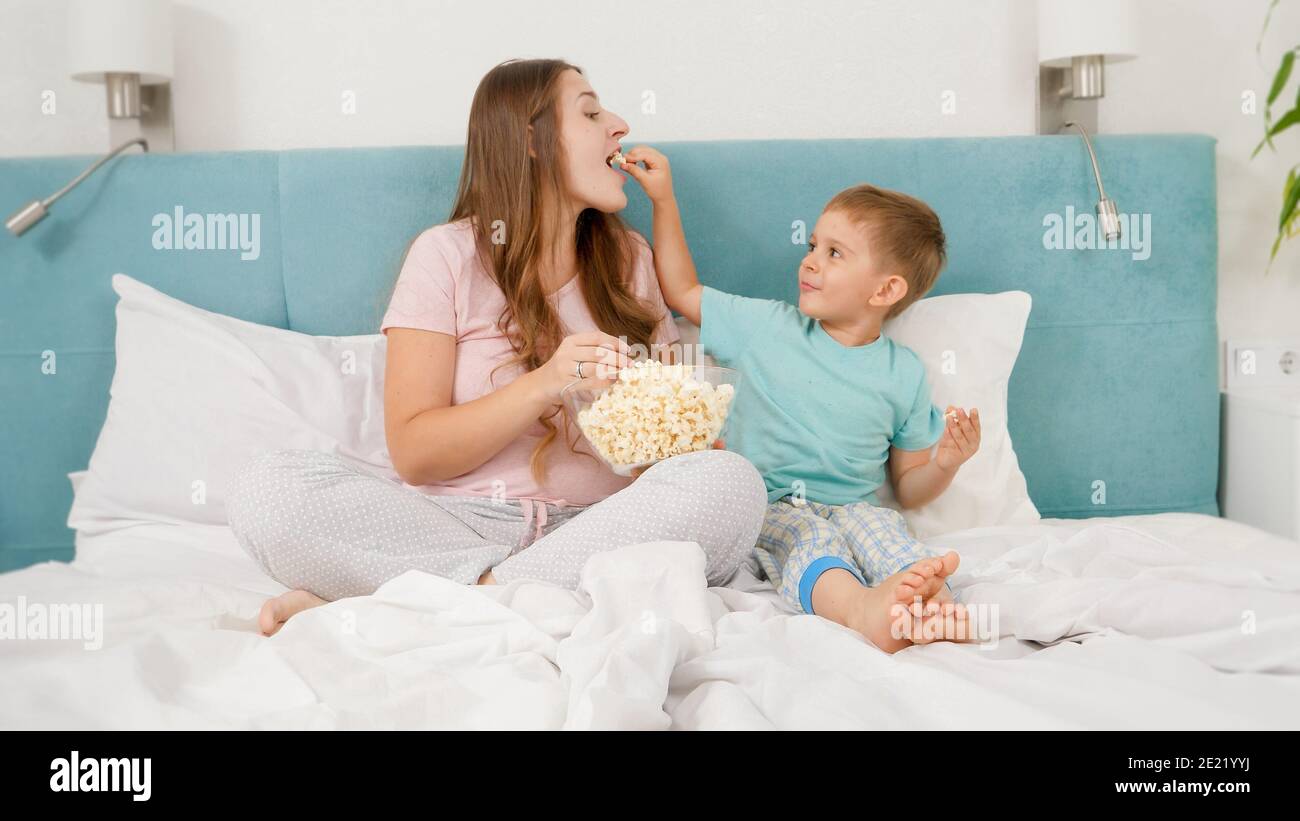 Kleiner Kleinkind Junge mit junger Mutter im Pyjama liegen im Bett am Wochenende und essen Popcorn aus großer Schüssel. Konzept der fröhlichen Kinder und Familie Stockfoto