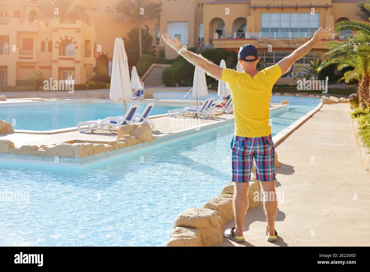 Glücklicher Mann hebt seine Hände in Hut und gelbem T-Shirt in der Nähe des Hotelpools hoch. Reisen, Sommerferien und Urlaubskonzept Stockfoto