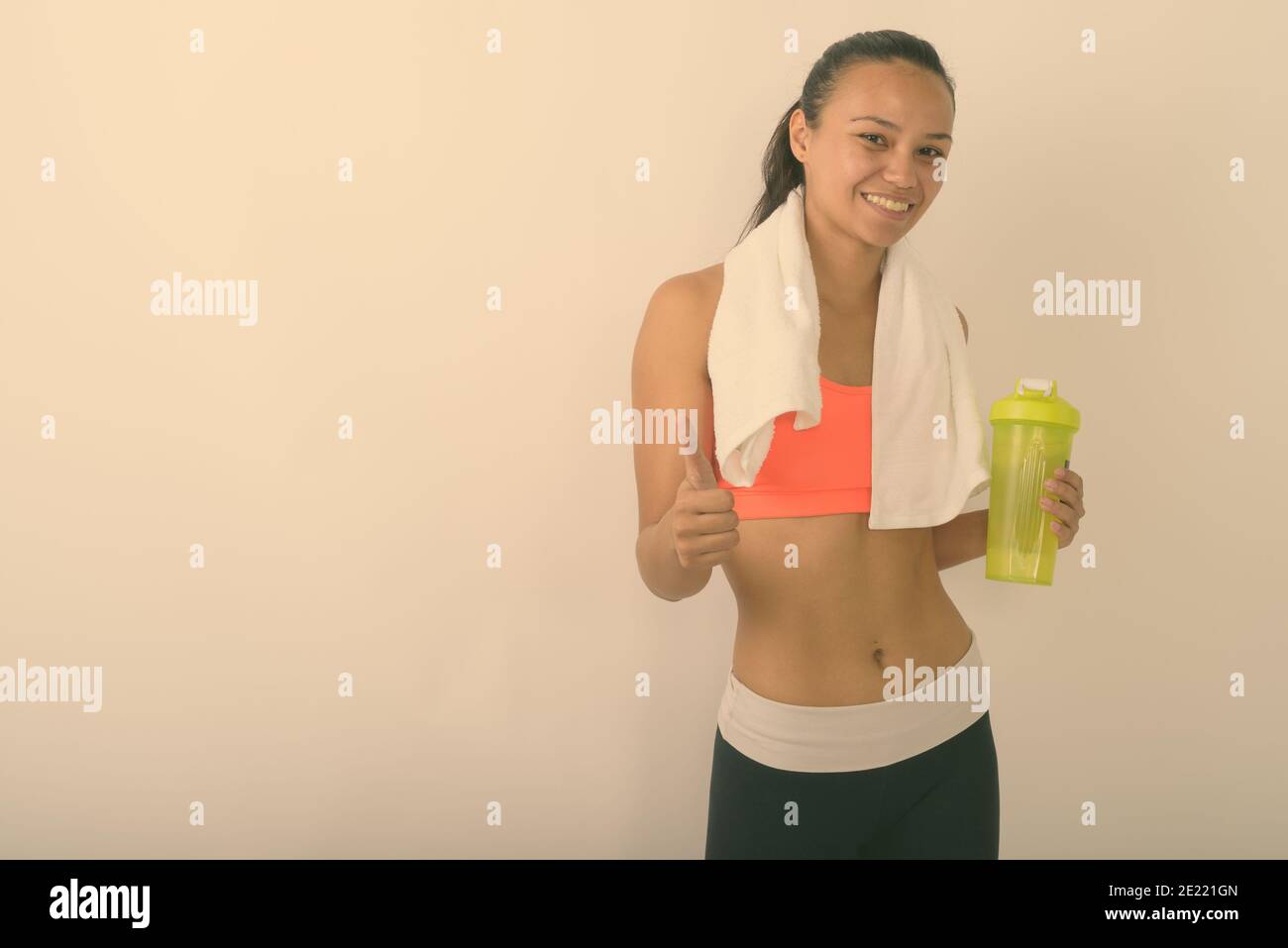 Studioaufnahme der jungen glücklichen asiatischen Frau lächelnd beim Geben Daumen hoch und halten Wasserflasche mit einem Handtuch um Hals bereit für das Fitnessstudio gegen Weiß Stockfoto