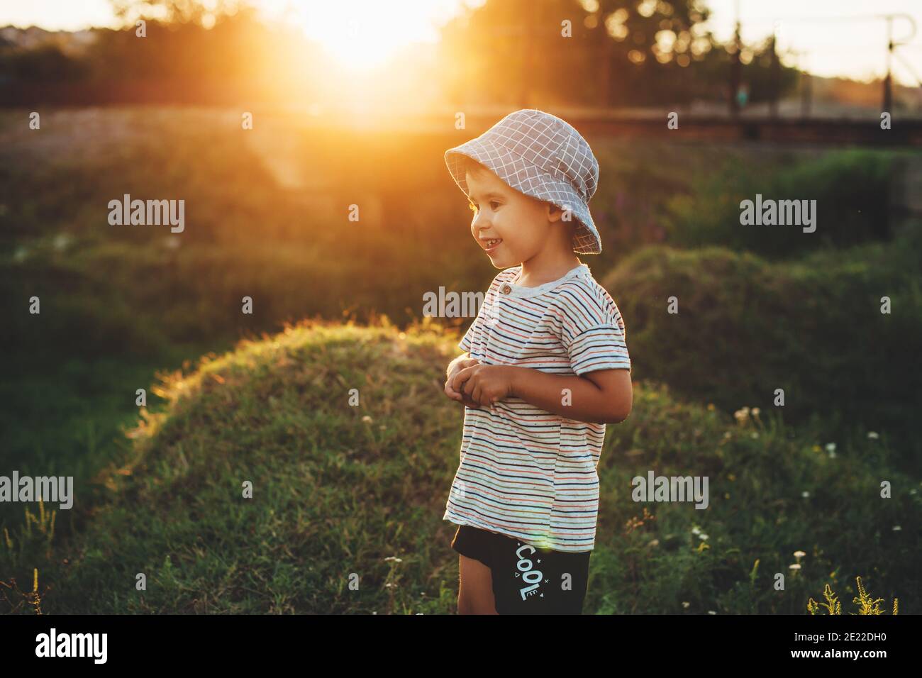 Charmanter Junge mit blauem Hut posiert in einem grünen Feld lächelt in der Sonne Stockfoto