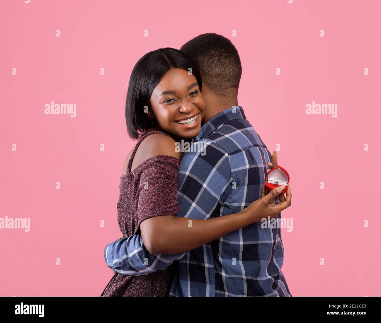 Glückliche schwarze Frau umarmt ihren Verlobten und hält Verlobungsring Auf pinkem Studiohintergrund Stockfoto