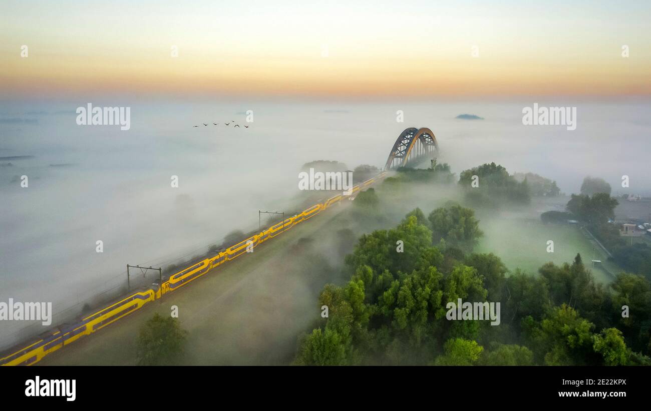 Zweidecker-Intercity-Zug von Nederlandse Spoorwegen durch dichten Nebel in Richtung Eisenbahnbogenbrücke, Gelderland, Niederlande Stockfoto