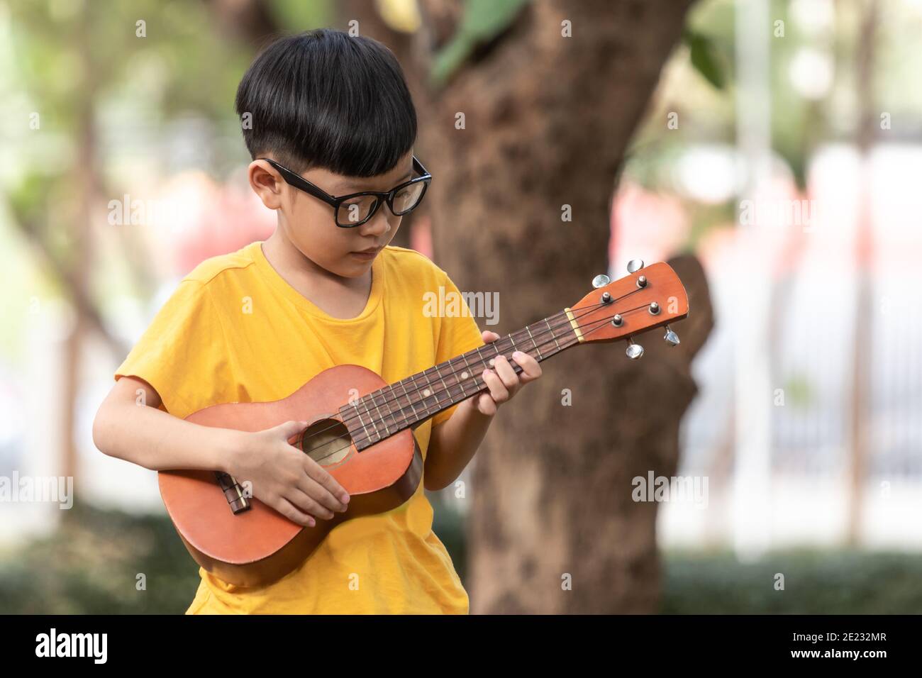 Ein asiatischer kleiner Junge mit Brille spielt glücklich die Ukulele. Asiatisches kleines Kind versucht, die Ukulele mit einem voll glücklichen Moment zu spielen. Stockfoto