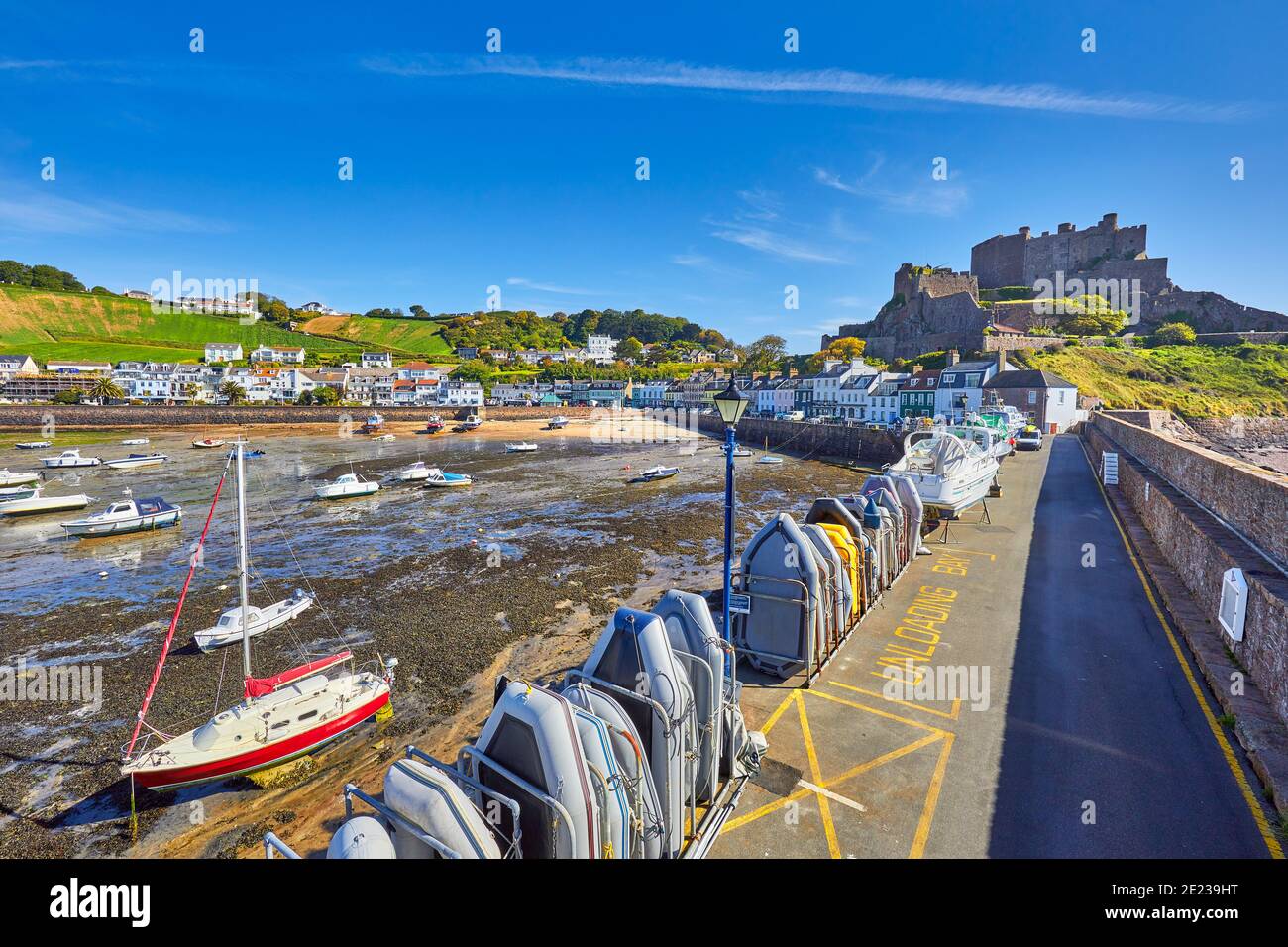 Bild von Gorey Harbour bei Ebbe mit geerdeten Booten, der Pier Bullworks und Gorey Castle im Hintergrund mit blauem Himmel. Jersey, Kanalinseln, Stockfoto