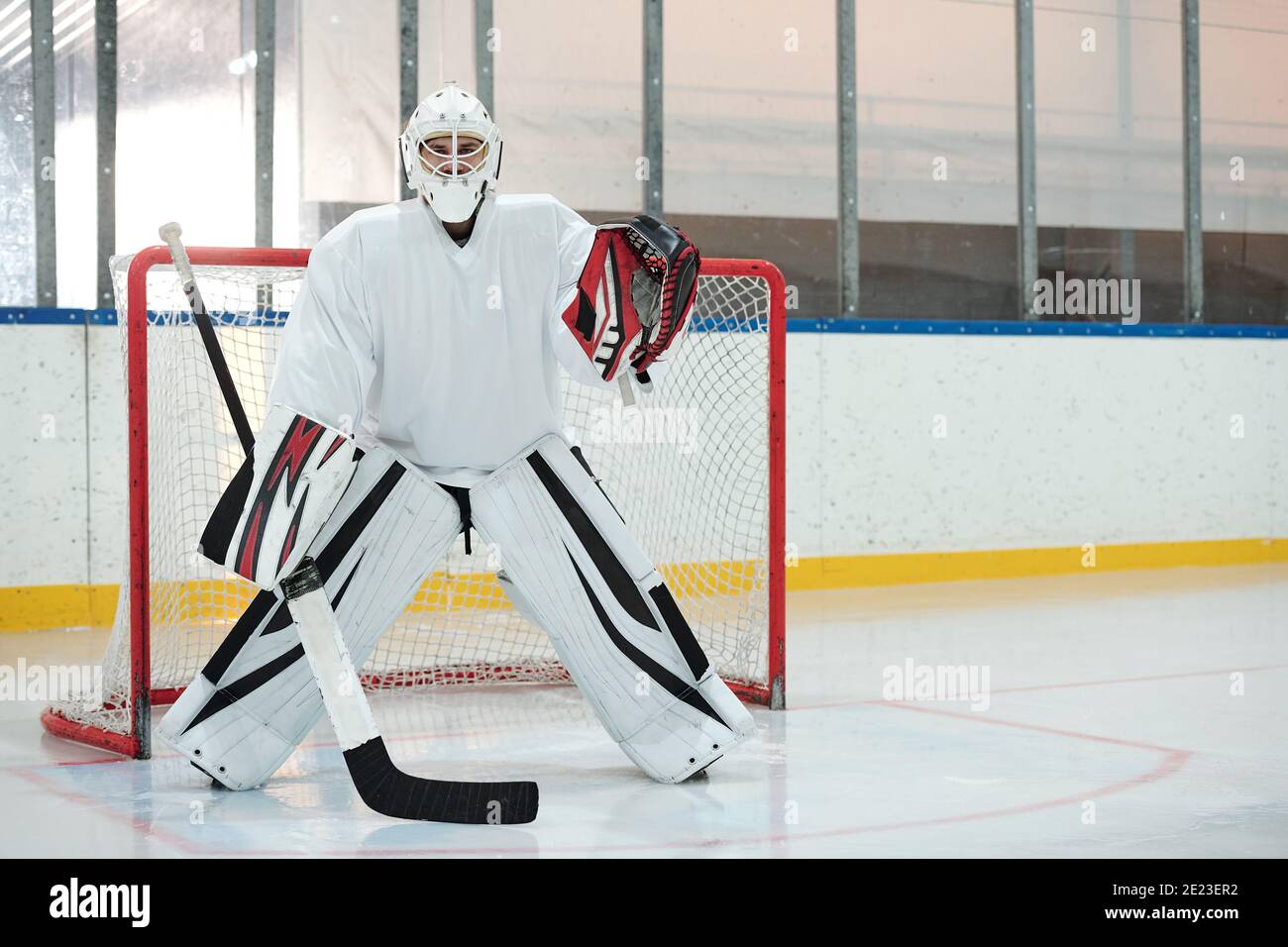 Hockeyspieler in weißer Sportuniform, Schutzhelm und Handschuhe halten Stock, während auf der Eisbahn gegen Netz stehen und warten auf Puck Stockfoto