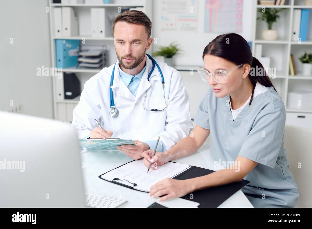 Zwei junge Kliniker in Uniform sitzen vor dem Schreibtisch Von Computermonitor und Beratung mit erfahrenen Kollegen in Video-Chat Stockfoto