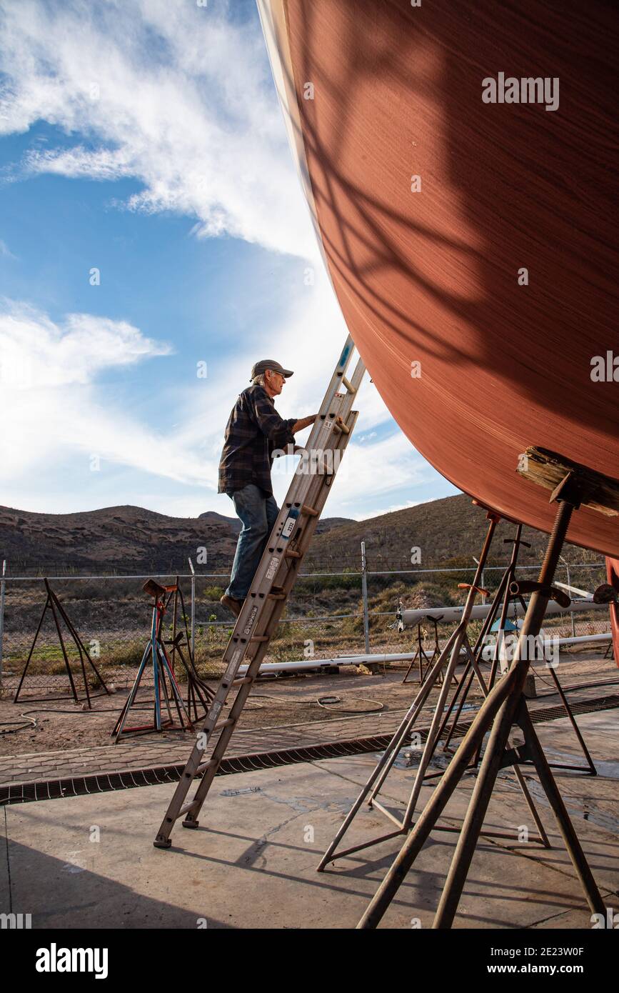 Ein älterer Mann klettert eine hohe Leiter, die sich an die Seite seines Segelbootes lehnt, auf den Bootsständen im Arbeitshof. Stockfoto