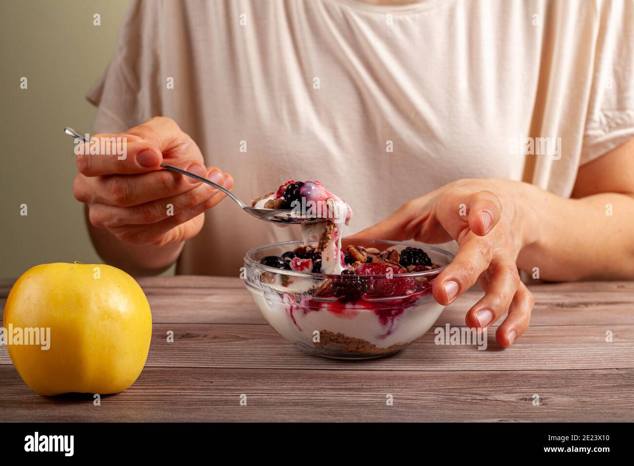 Eine kaukasische Frau isst eine frische, hausgemachte Glasschüssel mit cremigem Joghurt-Parfait mit Beeren, Müsli und Samen darin auf einem Holztisch Close Up Photo Stockfoto