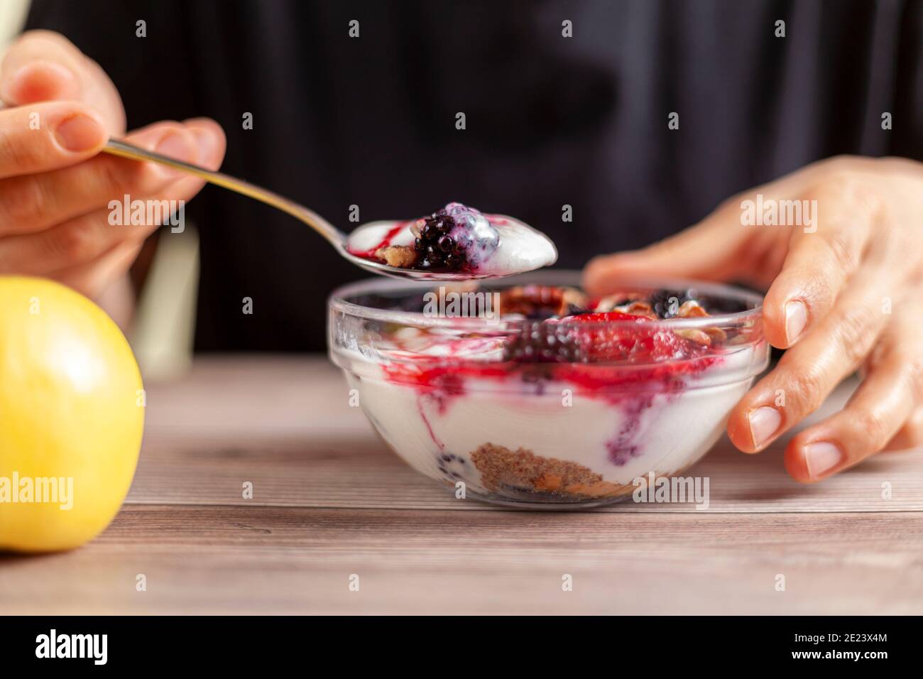 Frau trägt schwarzes Hemd isst eine frische hausgemachte Glasschüssel aus cremigem Joghurt-Parfait mit Beeren, Müsli und Samen darin auf Holztisch Nahansicht Foto Stockfoto