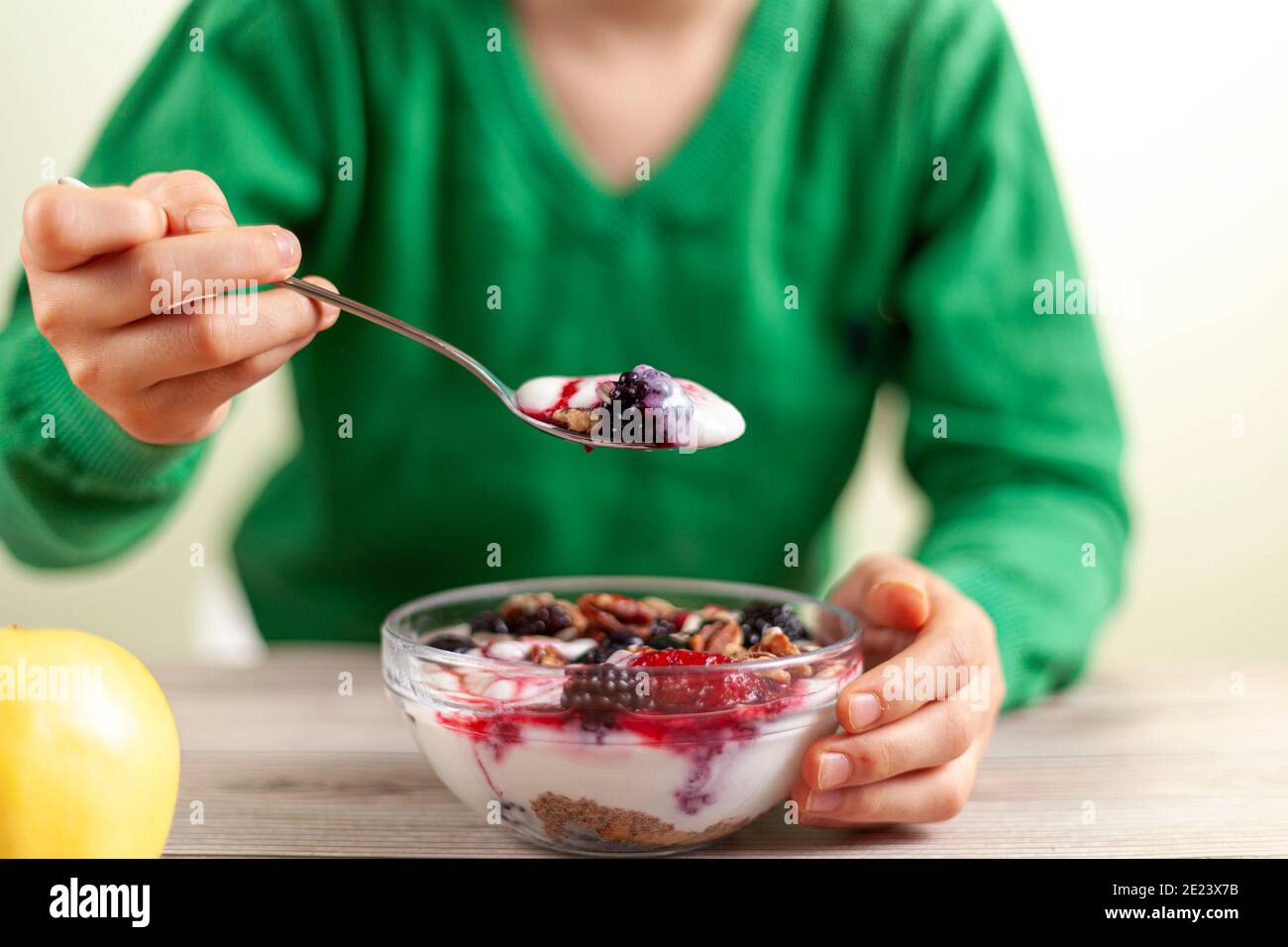 Ein Kind mit grünem Hemd isst frisch hausgemachte Glasschüssel mit cremigem Joghurt-Parfait mit Beeren, Müsli und Samen darin auf Holztisch Nahansicht Foto Stockfoto