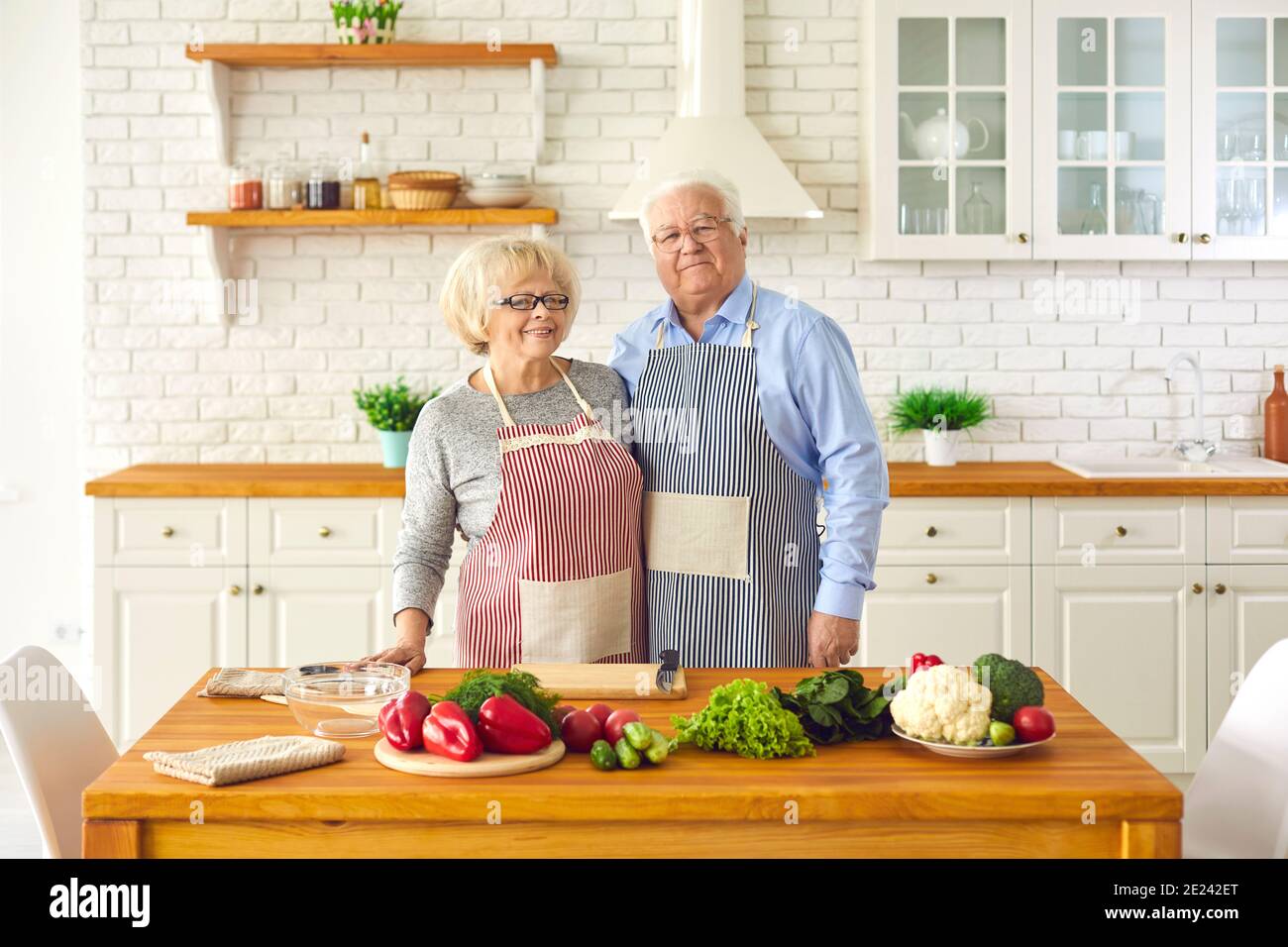 Seniorenpaar steht in Schürzen in der Küche in der Nähe von Kochprodukten und schaut auf die Kamera. Stockfoto