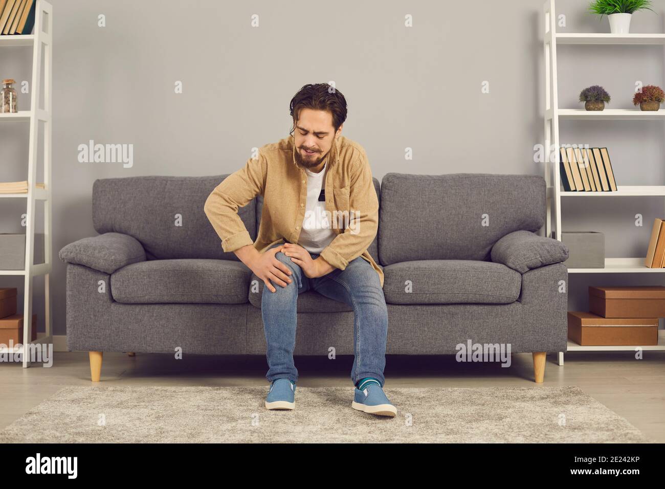 Junger Mann, der zu Hause auf der Couch sitzt und seine berührt Schmerzen im Knie mit Grimasse von Schmerzen Stockfoto
