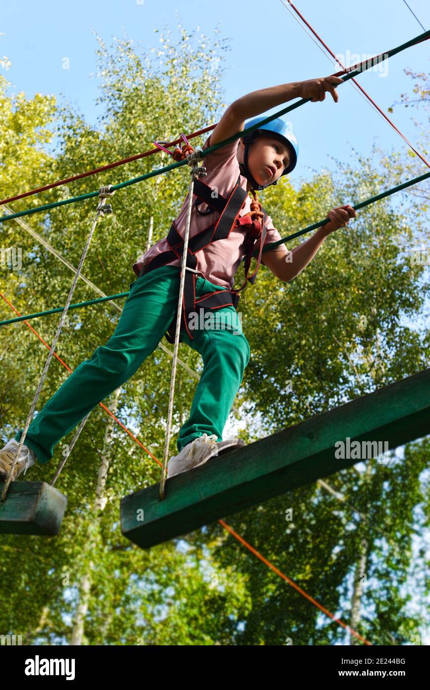 Ein Junge in einem Helm auf dem Kopf überwindet Hindernisse im Seilpark. Der Teenager bewegt sich auf Hängebalken mit einem Sicherheitsnetz. Stockfoto