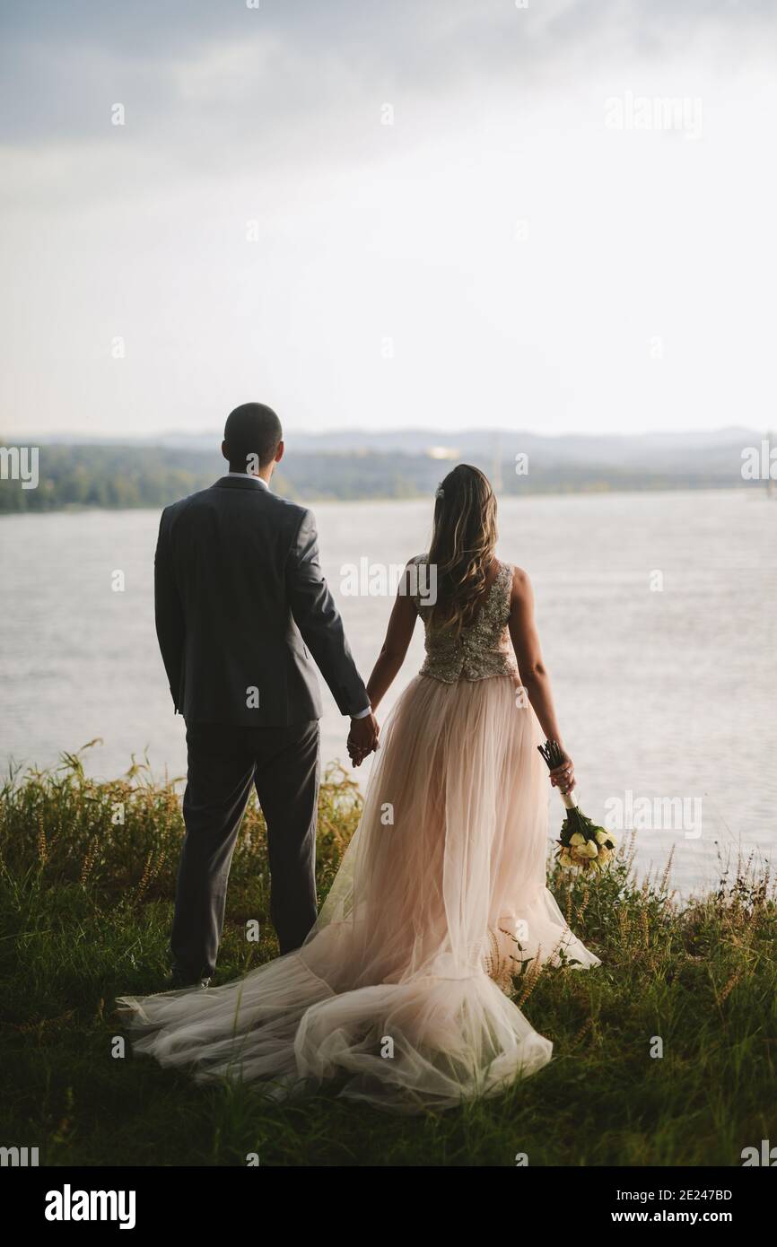 Gerade verheiratetes Paar, das in der Natur die Hände hält und einen schönen Blick auf den Fluss hat. Genießen Sie jeden Moment an ihrem besonderen Tag. Stockfoto