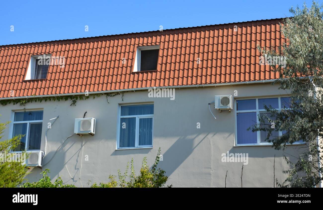 Ein kleines Wohnhaus mit Mansardendach aus rotem Lehm und Dachboden oder Mansardenfenstern, Klimaanlage im Freien an einer stuckgrauen Wand. Stockfoto
