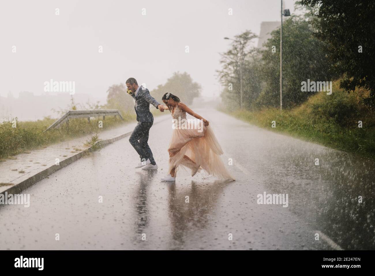 Alberne junge gerade verheiratete Paar überqueren Straße an regnerischen Tag. Laufen in nasser zeremonieller Kleidung. Stockfoto