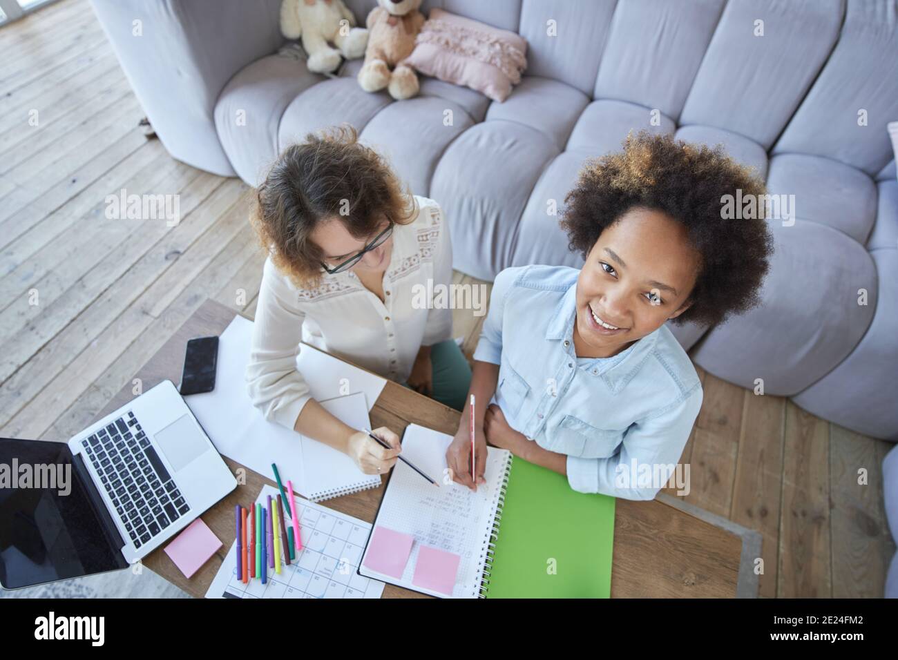 Fröhlich gemischt Rennen teen Mädchen lächeln bei der Kamera während tun Hausaufgaben zusammen mit Lehrerin im Wohnzimmer bei Zu Hause Stockfoto