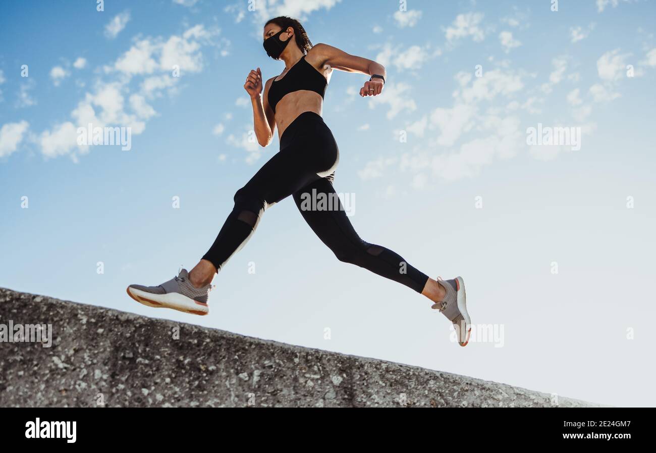Low-Angle-Aufnahme einer sportlichen Sportlerin, die beim Laufen über einer Wand gegen den blauen Himmel trainiert. Frau in Sportbekleidung und Schutzmaske Ausübung o Stockfoto